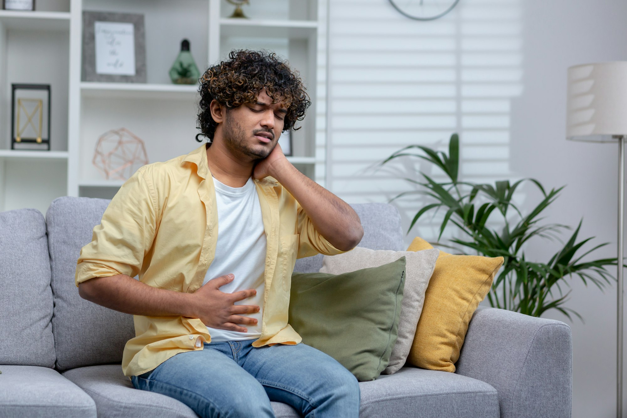 Young man sick at home sitting on sofa, Latin American has severe stomach pain and nausea, poisoned